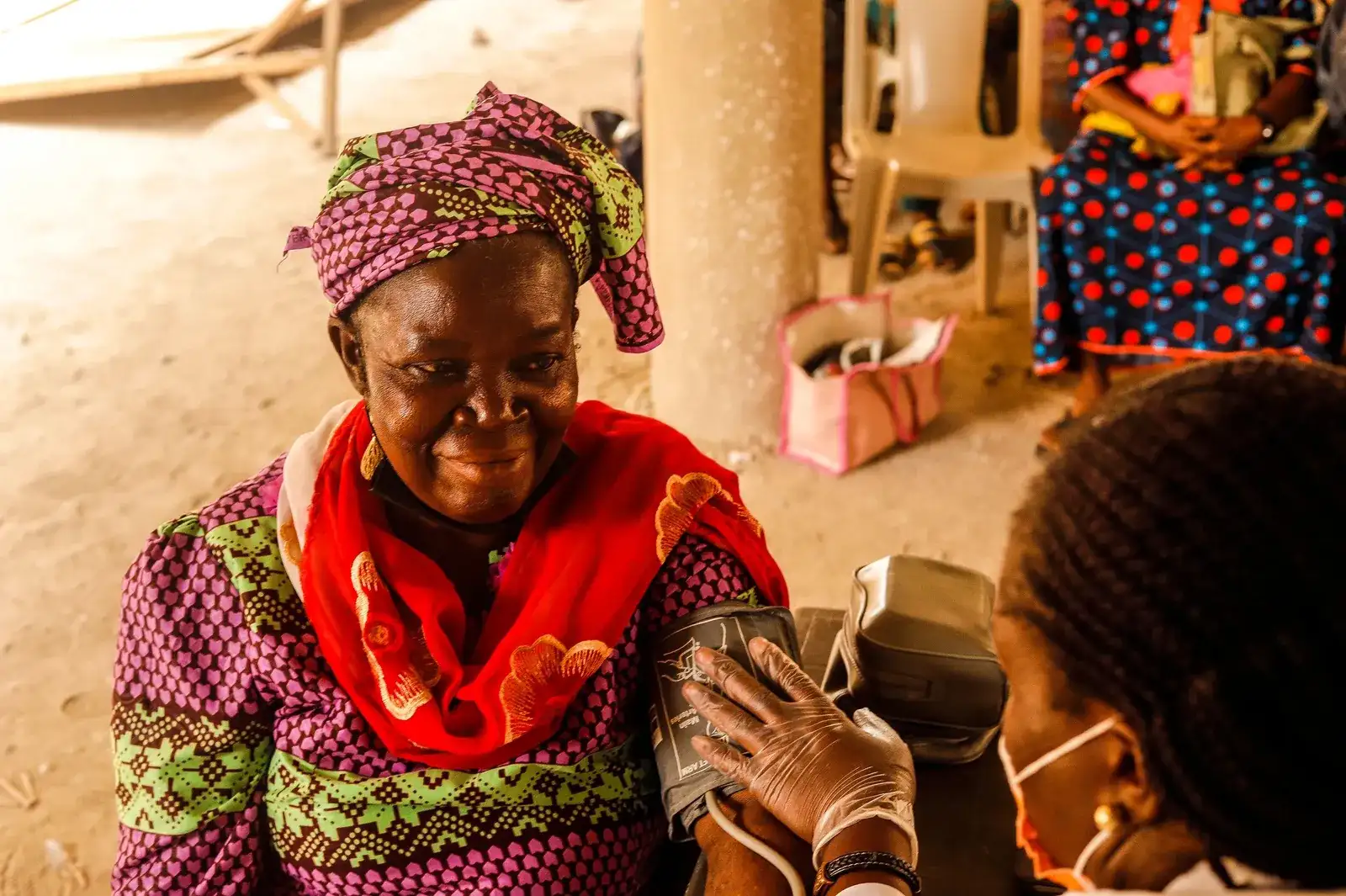 A smiling older woman in a headwrap has her blood pressure taken by a gloved health worker