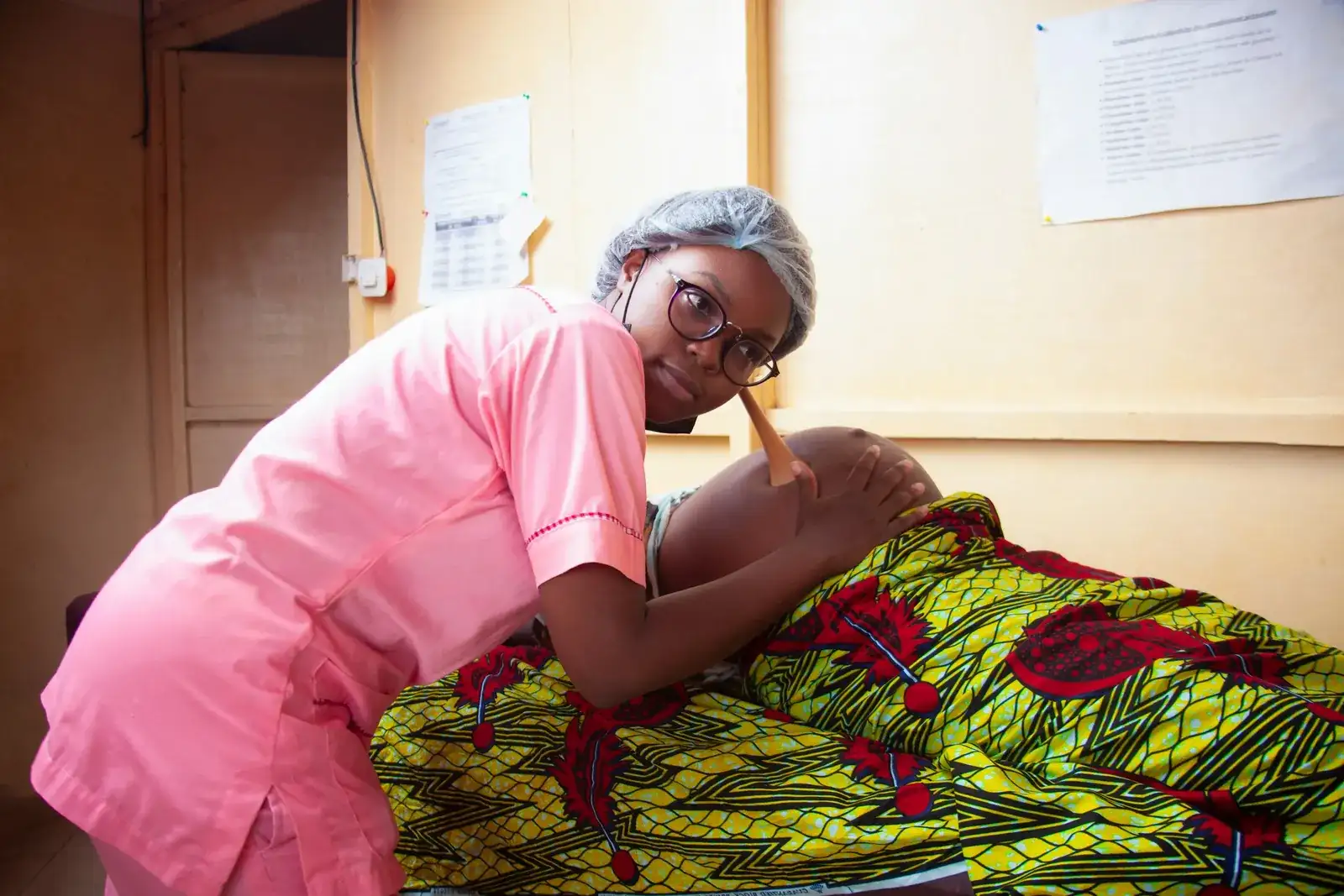 A health worker holds a fetal stethoscope to a pregnant woman's belly in a clinic